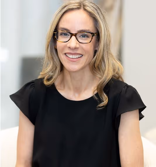 A woman with wavy blonde hair, wearing glasses and a black blouse, smiling while seated in an office setting.