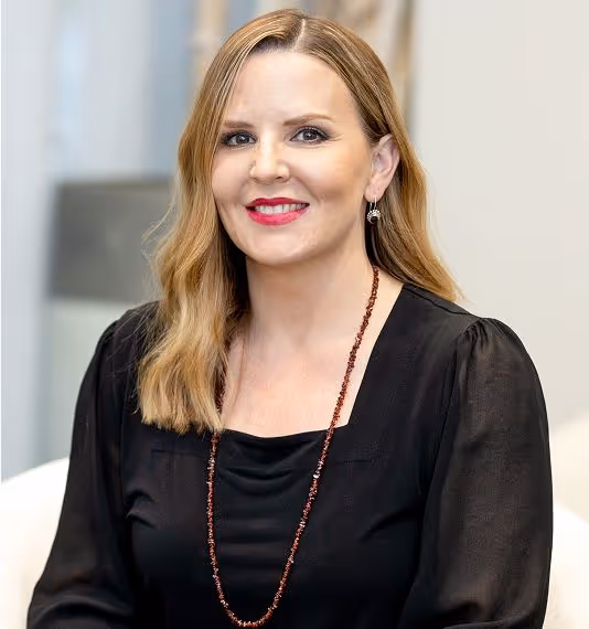 A woman with long brown hair and a black blouse smiling at the camera, wearing a red lipstick and a necklace.