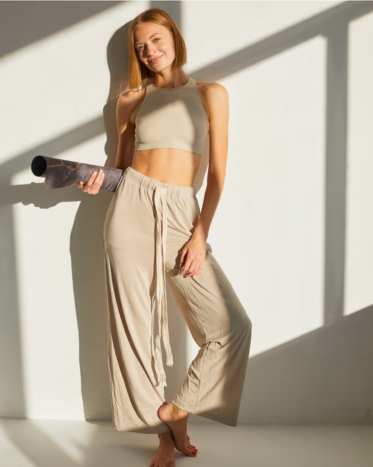 A woman in a beige crop top and loose beige pants, holding a yoga mat under her arm, standing in a relaxed pose with one foot slightly forward.