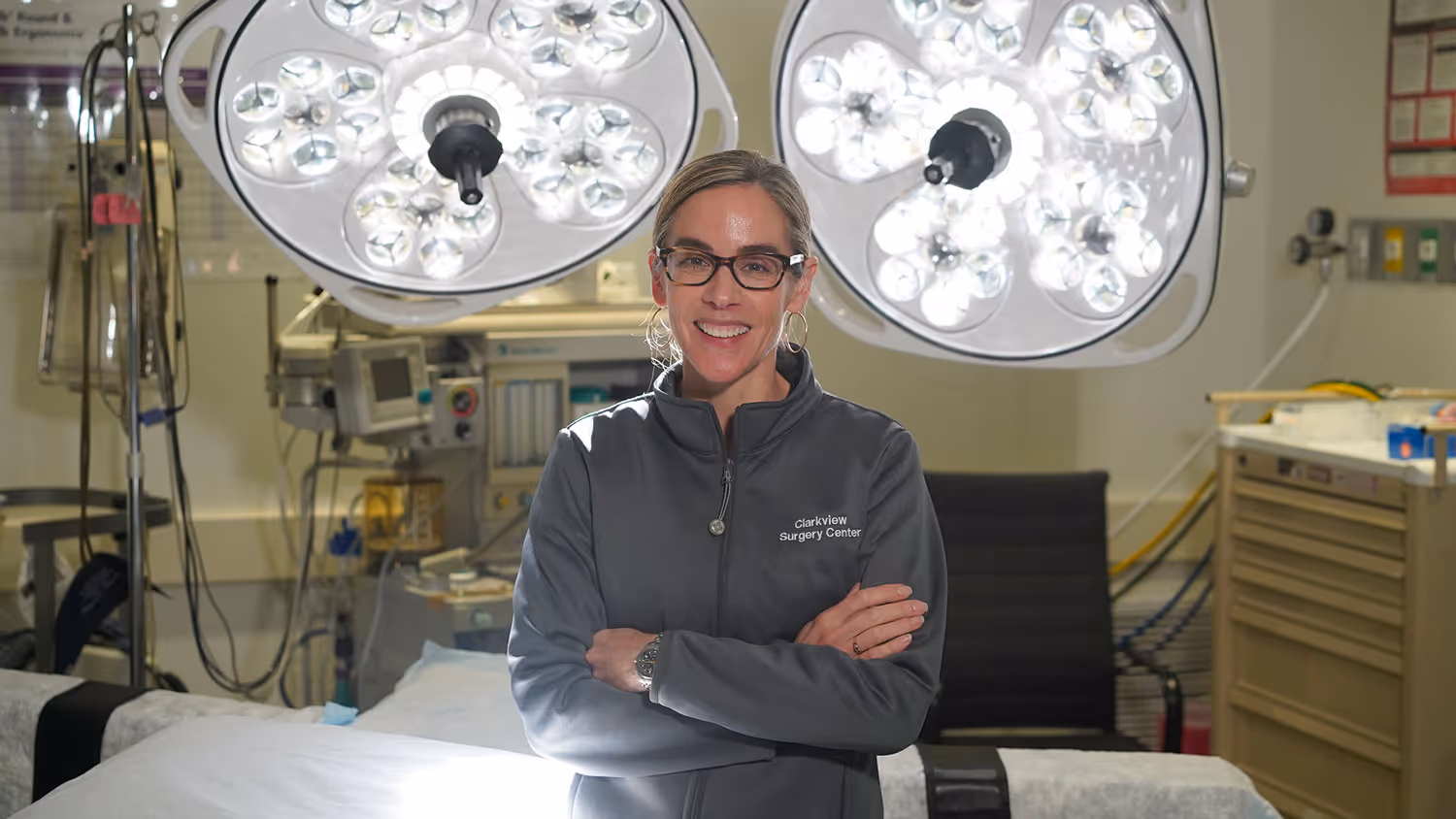 A woman with glasses smiles while standing in a medical environment, wearing a dark jacket with 'Caroline Surgery Center' text on the left chest, with overhead surgical lights in the background.