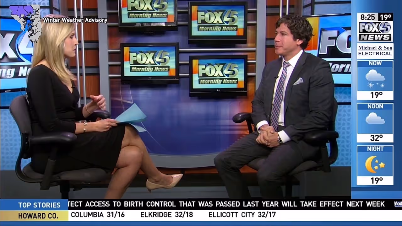 A televised news interview featuring two individuals, one woman with long blonde hair wearing a black dress and one man with dark hair wearing a light-colored suit and tie. They are seated facing each other, discussing a topic with visual elements on the.