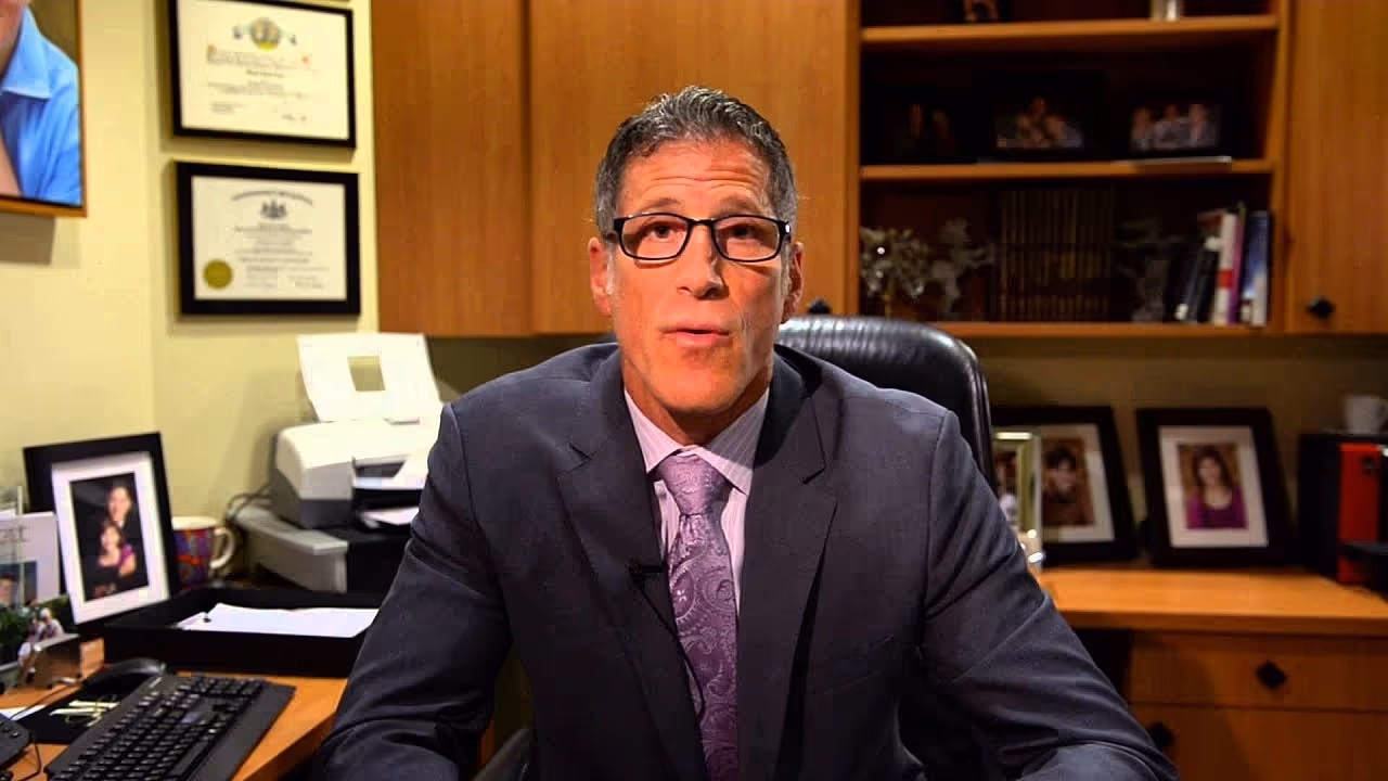 A man in a suit and glasses sitting at a desk with a concerned expression, in an office setting with framed certificates and photographs in the background.