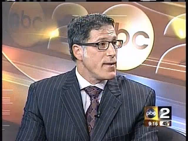 A man wearing glasses and a pinstriped suit, with a white shirt and a patterned tie, appears to be speaking during a television segment. The background features the ABC 2 logo and a stylized graphic design.