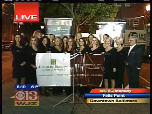 A group of women standing together in formal attire, holding a large banner with the text 'Cosmetic Surgery Center of Maryland' and 'Live' at the top left. Several promotional signs are visible in the background.