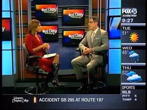 A woman in a red dress and black knee-high boots sitting on a stool while interviewing a man in a gray suit and glasses, who is also seated on a stool. They are discussing a topic related to cosmetic surgery, with a weather and news ticker visible on the.