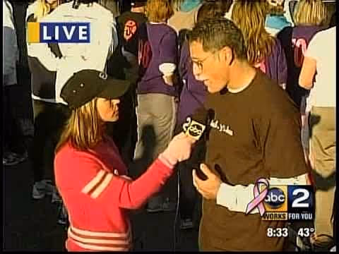 A woman wearing a black cap and pink long-sleeve shirt is interviewing a man in a brown t-shirt. The man is speaking while the woman holds a microphone towards him. In the background, a crowd of people is partially visible.