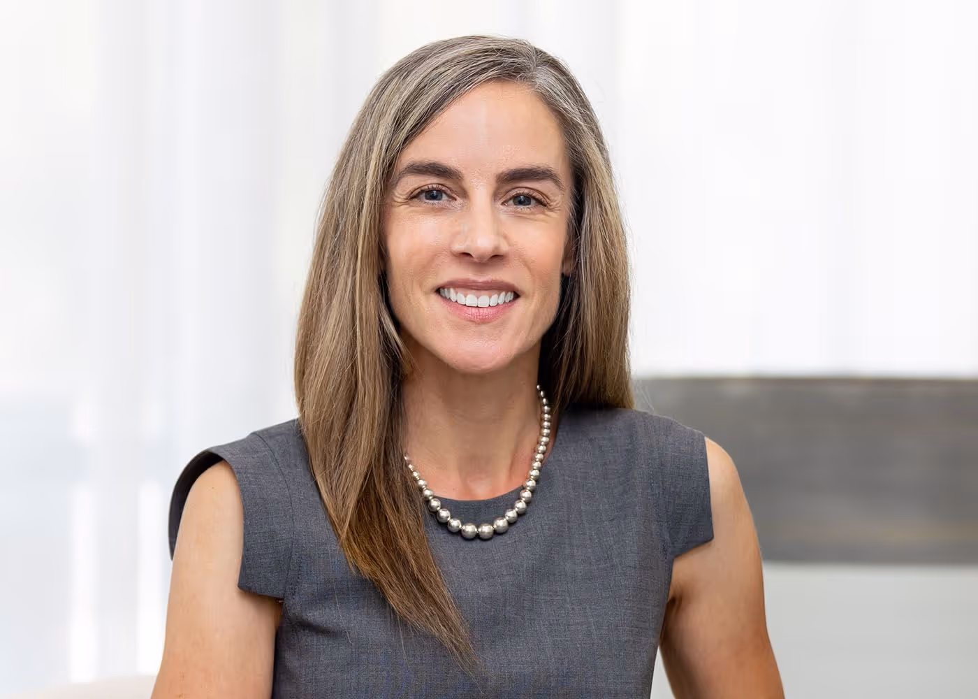 A woman with long, straight brown hair wearing a gray sleeveless dress and a pearl necklace, smiling directly at the camera.