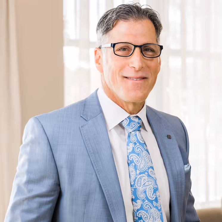 A man dressed in a light blue suit with a paisley tie, wearing glasses, and smiling at the camera.
