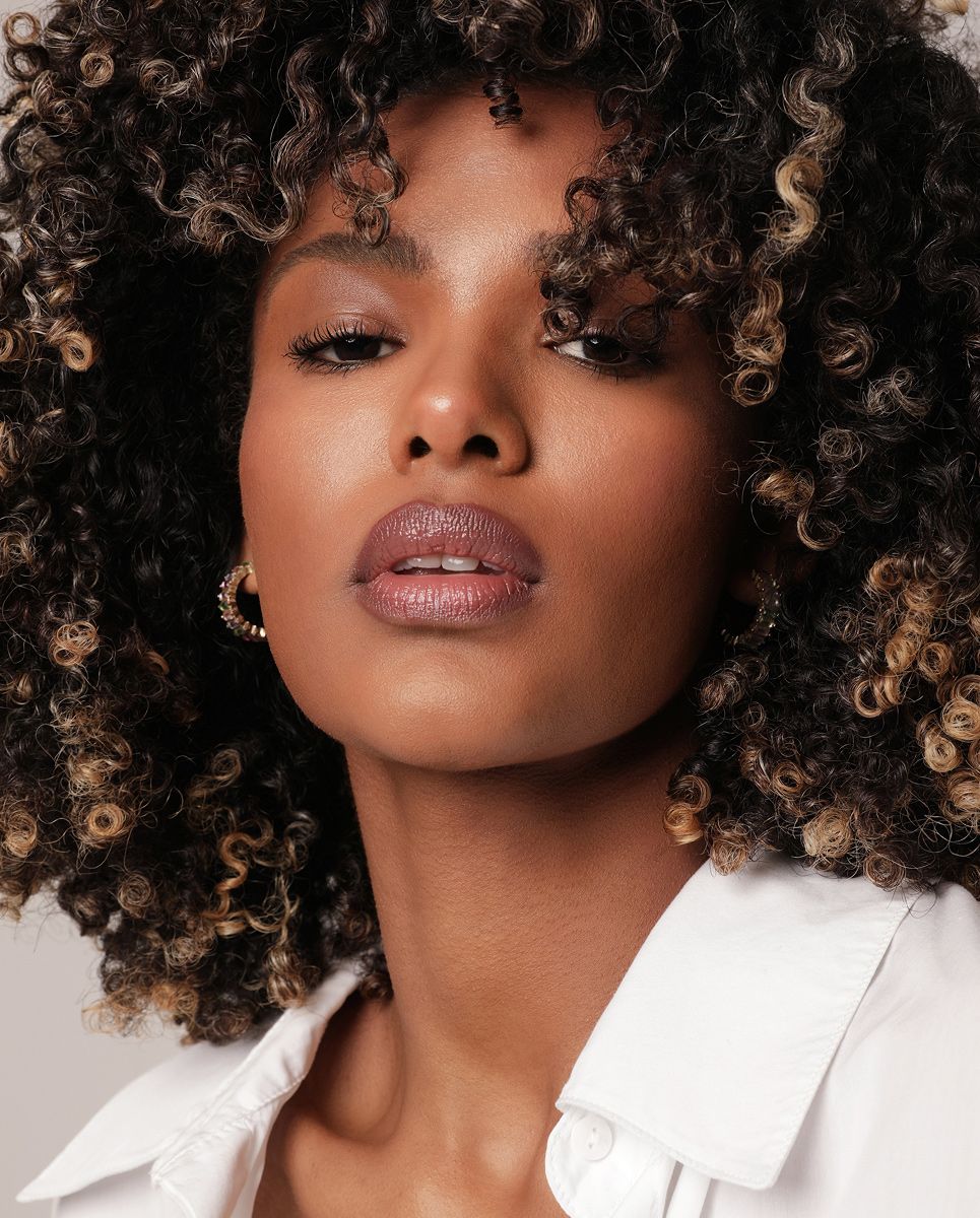 A close-up image of a woman with curly hair, wearing hoop earrings, and showcasing a natural makeup look with a focus on her lips.