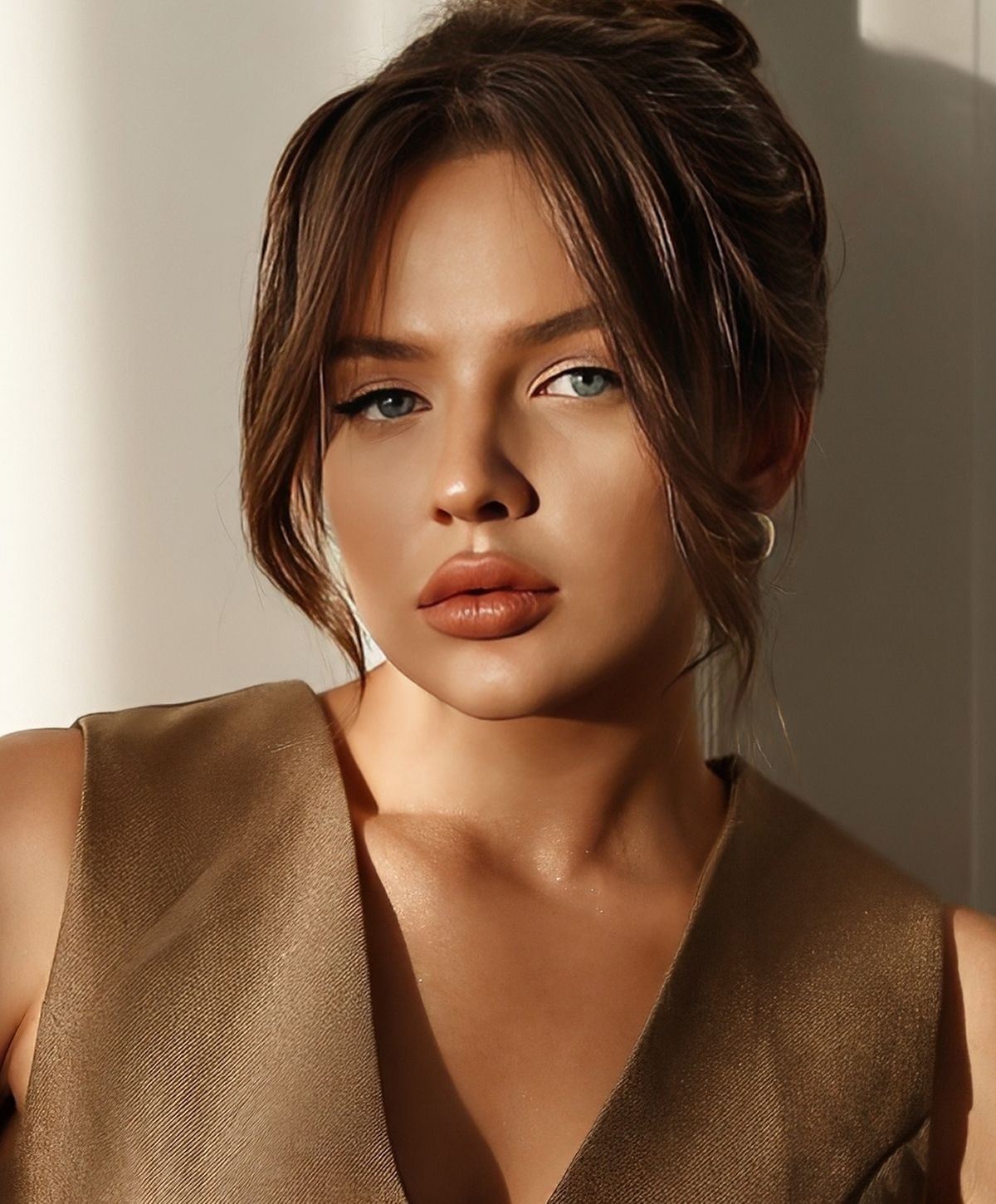 A woman with long brown hair styled in a bun, wearing a sleeveless bronze top, has a neutral expression. She has blue eyes and is positioned to the side, with soft lighting highlighting her facial features.