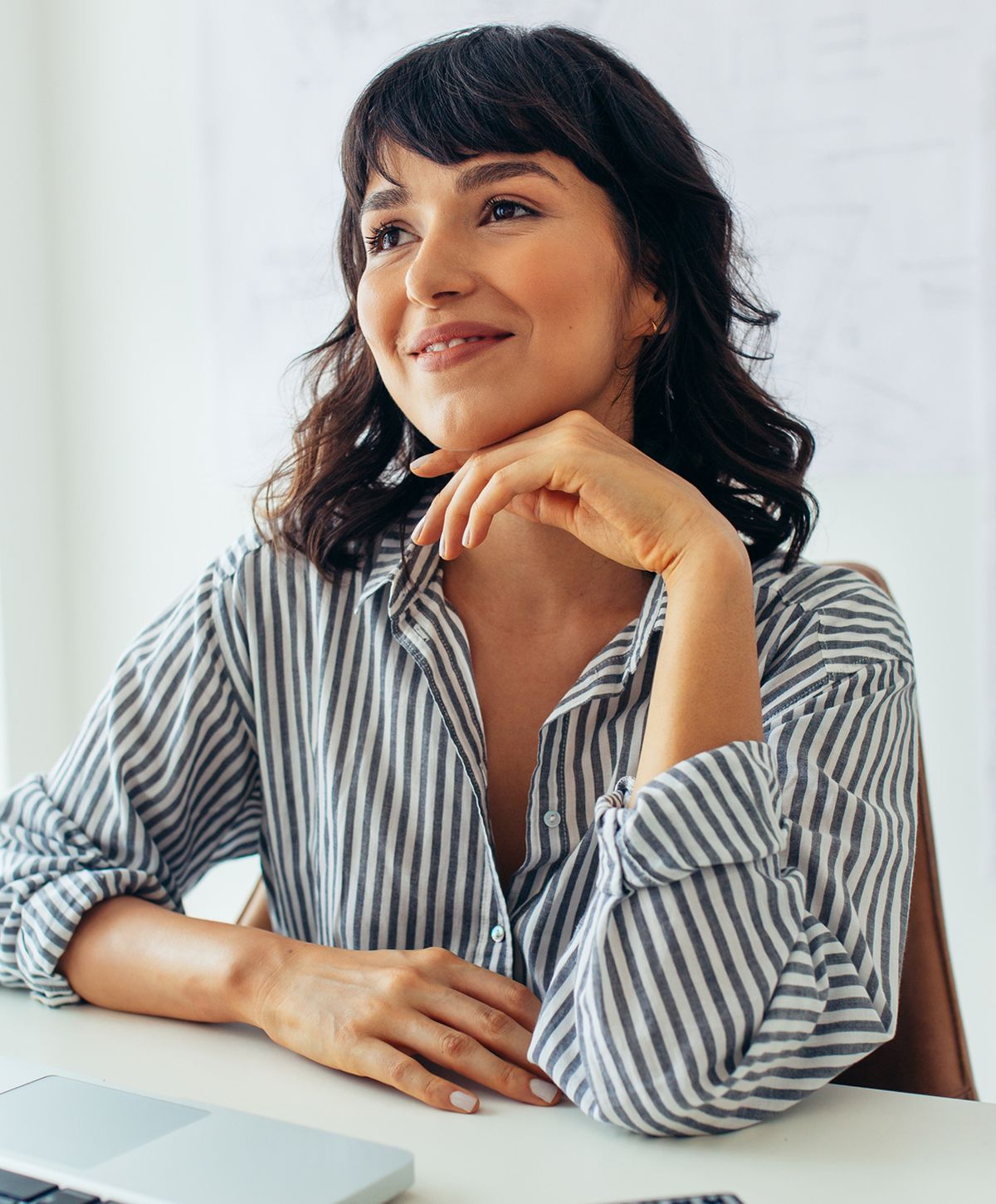 A woman with medium-length dark hair styled in loose waves, wearing a striped button-up shirt, sitting at a table with a thoughtful expression, resting her chin on her hand.