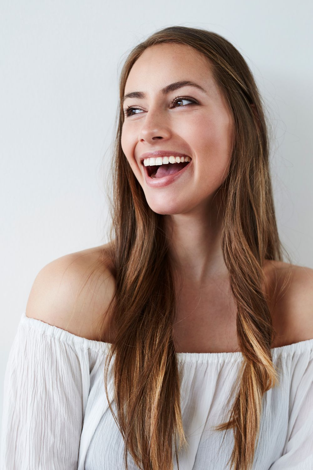 A woman with long, wavy brown hair and a bright smile, wearing an off-the-shoulder white top.
