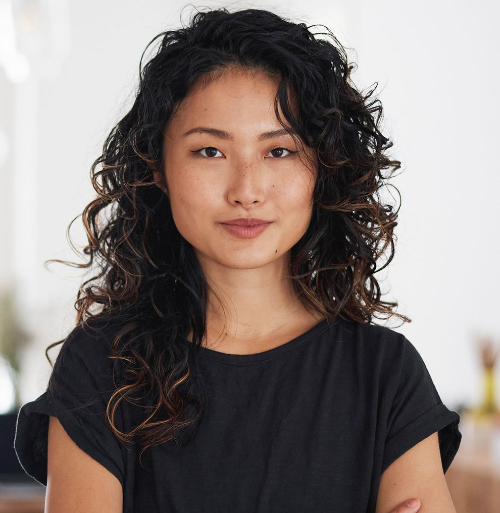 A woman with curly hair stands confidently, arms crossed, wearing a black shirt. She has a subtle smile and an expression of determination.