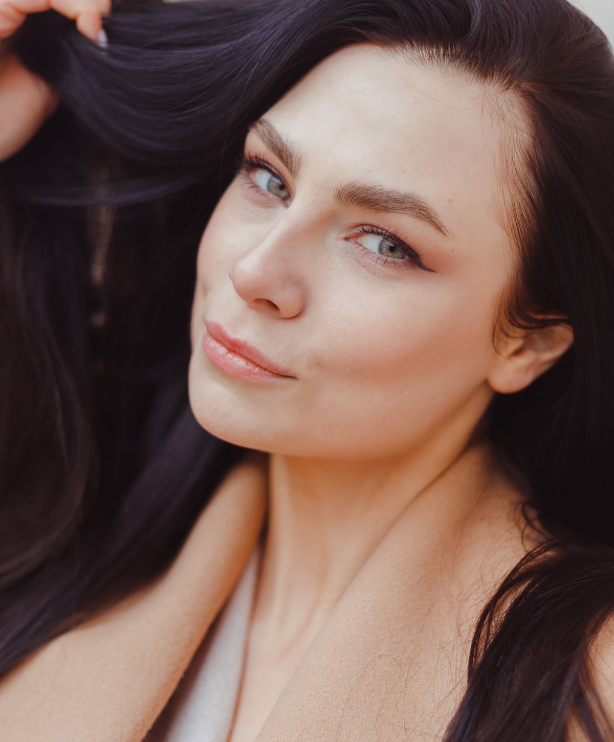 A woman with long dark hair, fair skin, and natural makeup is playfully holding her hair while looking directly at the camera with a slight smile.
