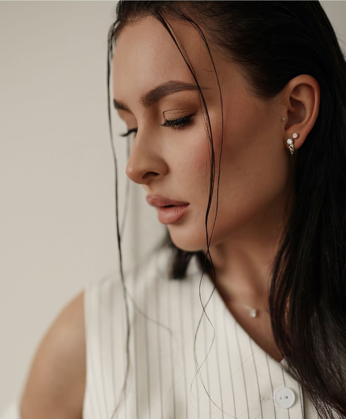 A woman with long dark hair styled down, wearing a white striped top, looking slightly to the side. She has subtle makeup and is wearing earrings.