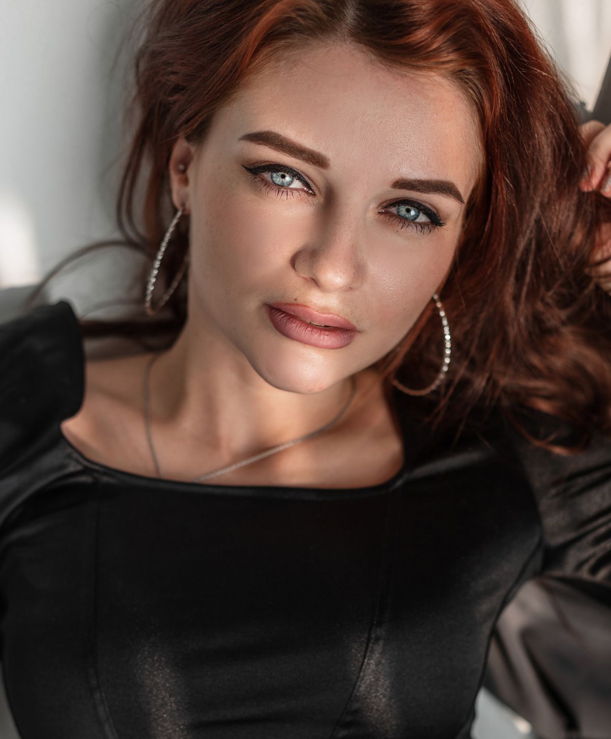 A woman with long, wavy red hair and blue eyes, wearing a black top, poses for the camera with a subtle smile and silver hoop earrings.