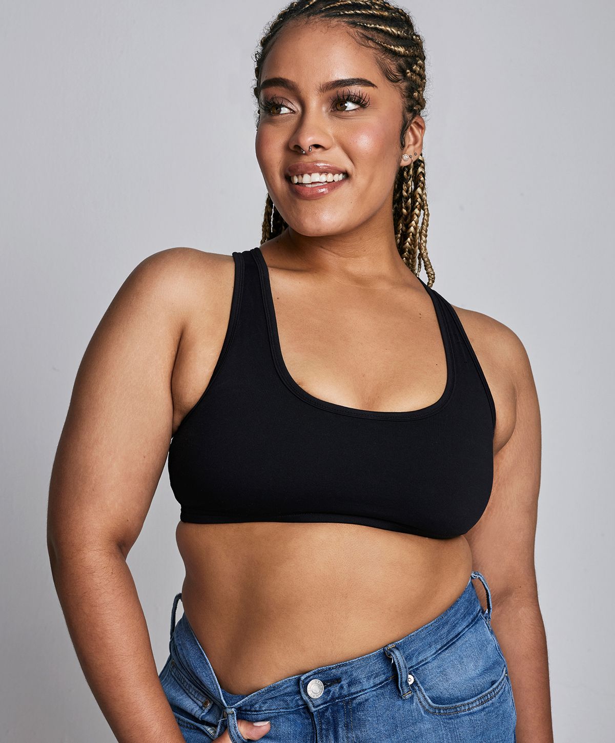 A woman with braided hair is smiling while wearing a black sports bra and blue jeans.