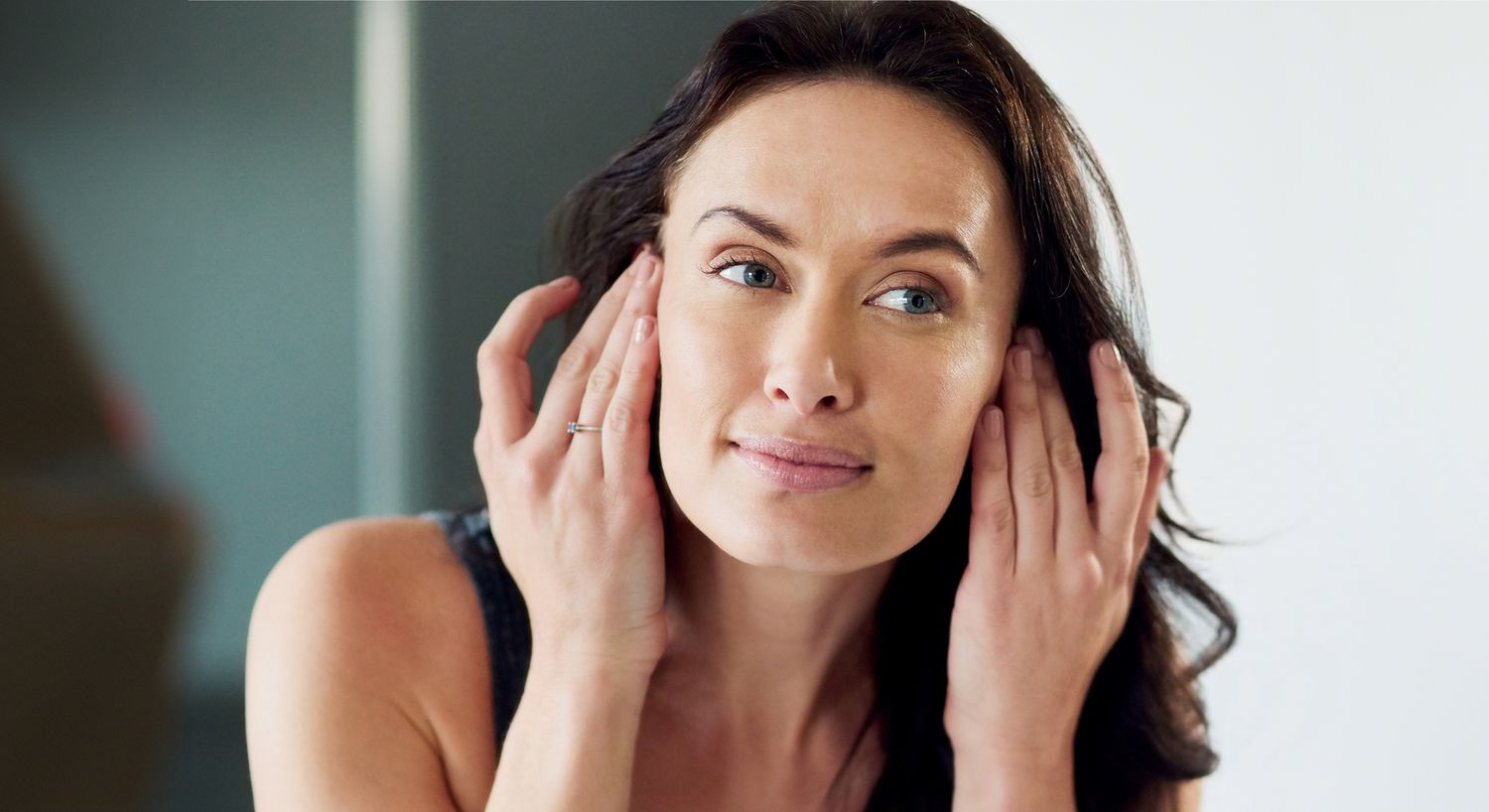 A woman gently touching her face, showing a thoughtful expression, with smooth skin and loose dark hair.