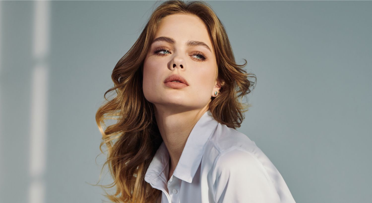 A woman with long, wavy hair wearing a white shirt, gazing slightly upward with a serene expression.