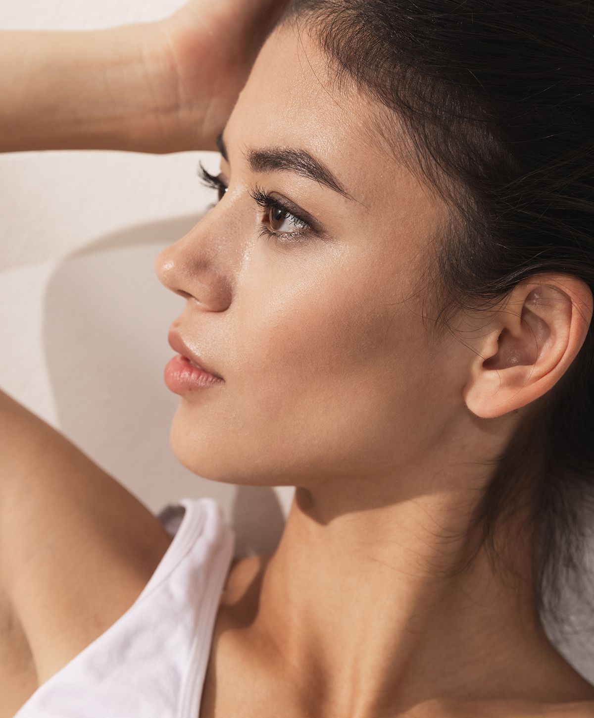 A side profile of a woman with long dark hair, lightly tanned skin, and natural makeup, gazing thoughtfully with her hand resting behind her head.