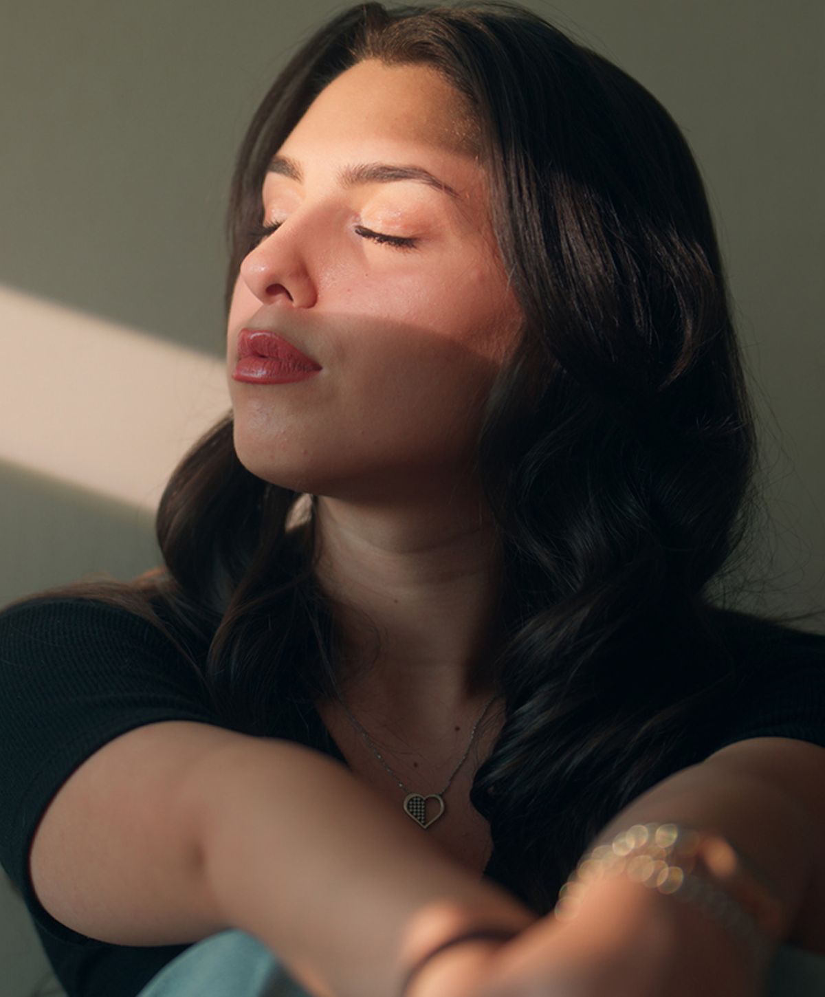 A woman with long dark hair, wearing a black top, appears to be enjoying sunlight with her eyes closed, and she has a heart-shaped pendant necklace.
