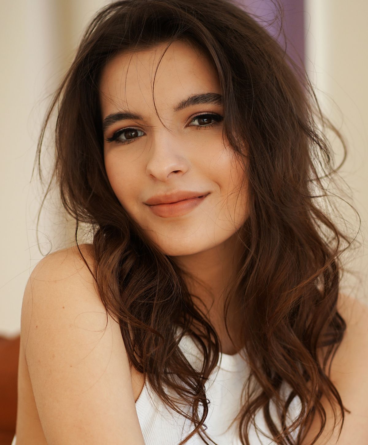 A smiling young woman with long, wavy brown hair, wearing a white sleeveless top, looking directly at the camera.
