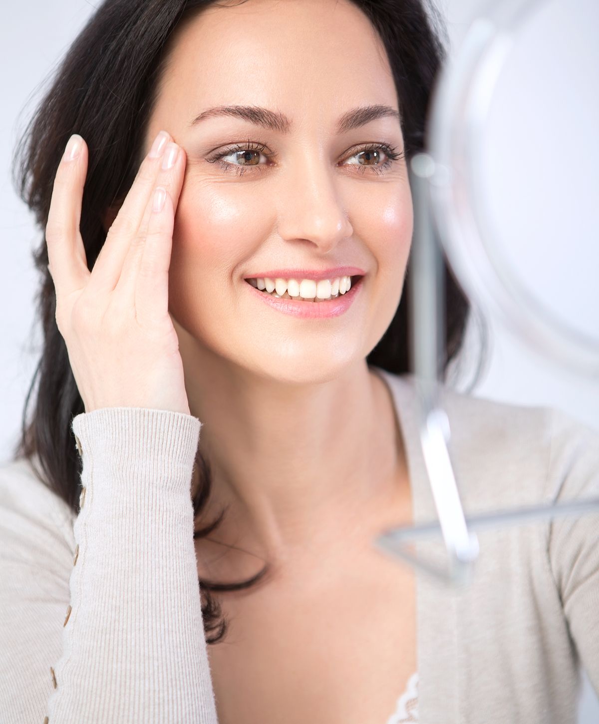 A woman with long dark hair smiling while touching her cheek, looking at her reflection in a handheld mirror.