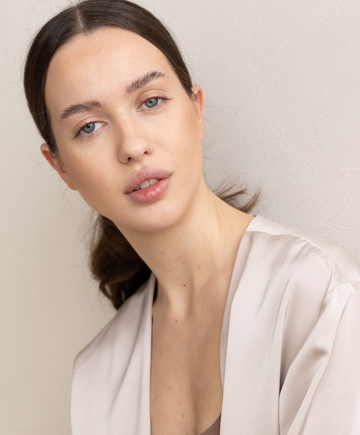 A woman with long brown hair styled in a loose ponytail, wearing a light-colored blouse, looking directly at the camera with a neutral expression.