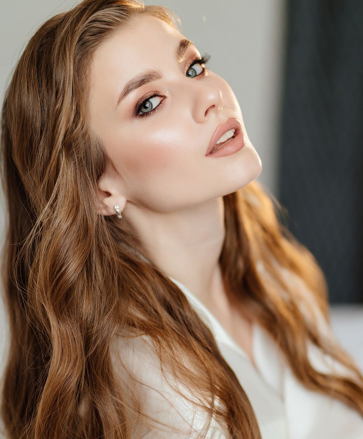 A woman with long, wavy brown hair and subtle makeup, looking over her shoulder with a soft expression.