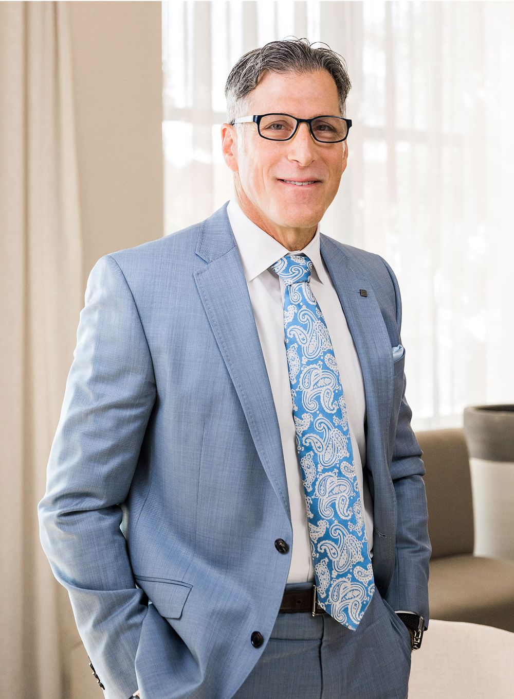 A man in a light blue suit and white shirt, wearing black glasses and a blue paisley tie, stands smiling with hands in pockets in a well-lit interior setting.