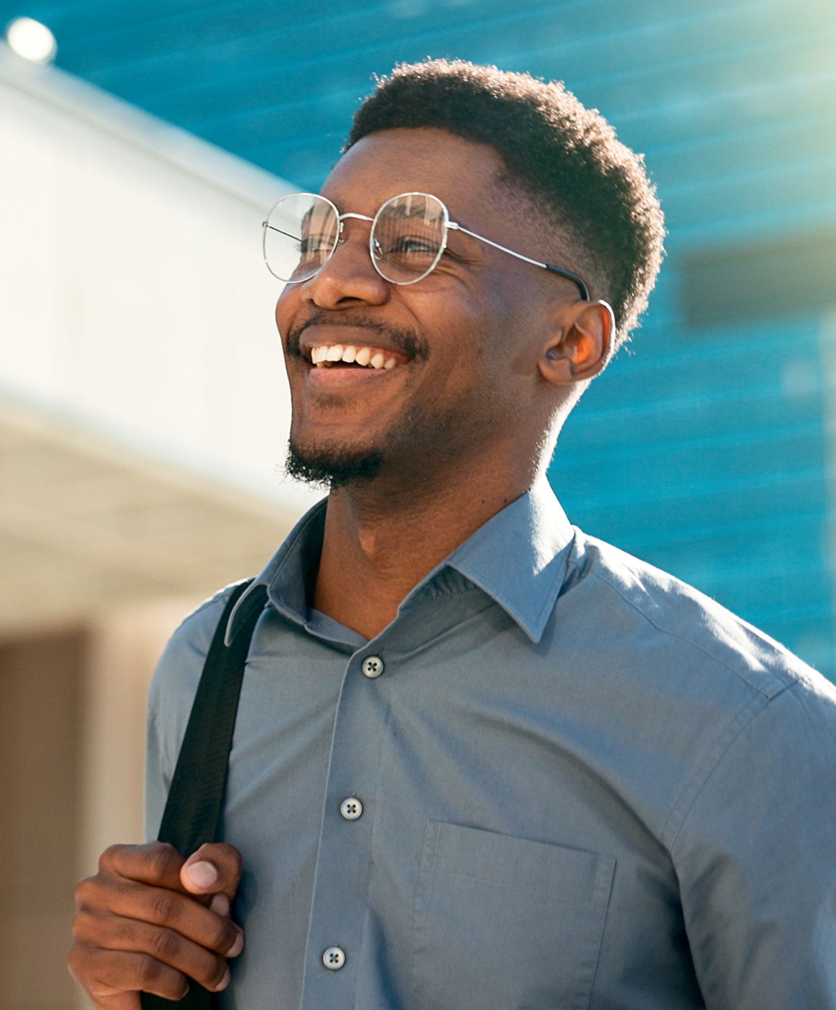 A man wearing glasses and a light blue shirt, smiling while carrying a shoulder bag.