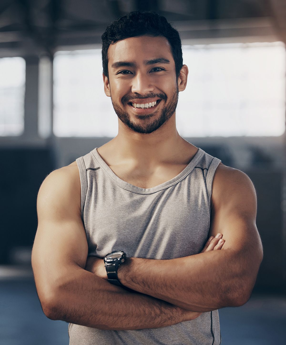 A smiling man with short dark hair and a beard, wearing a gray sleeveless shirt, standing with arms crossed.