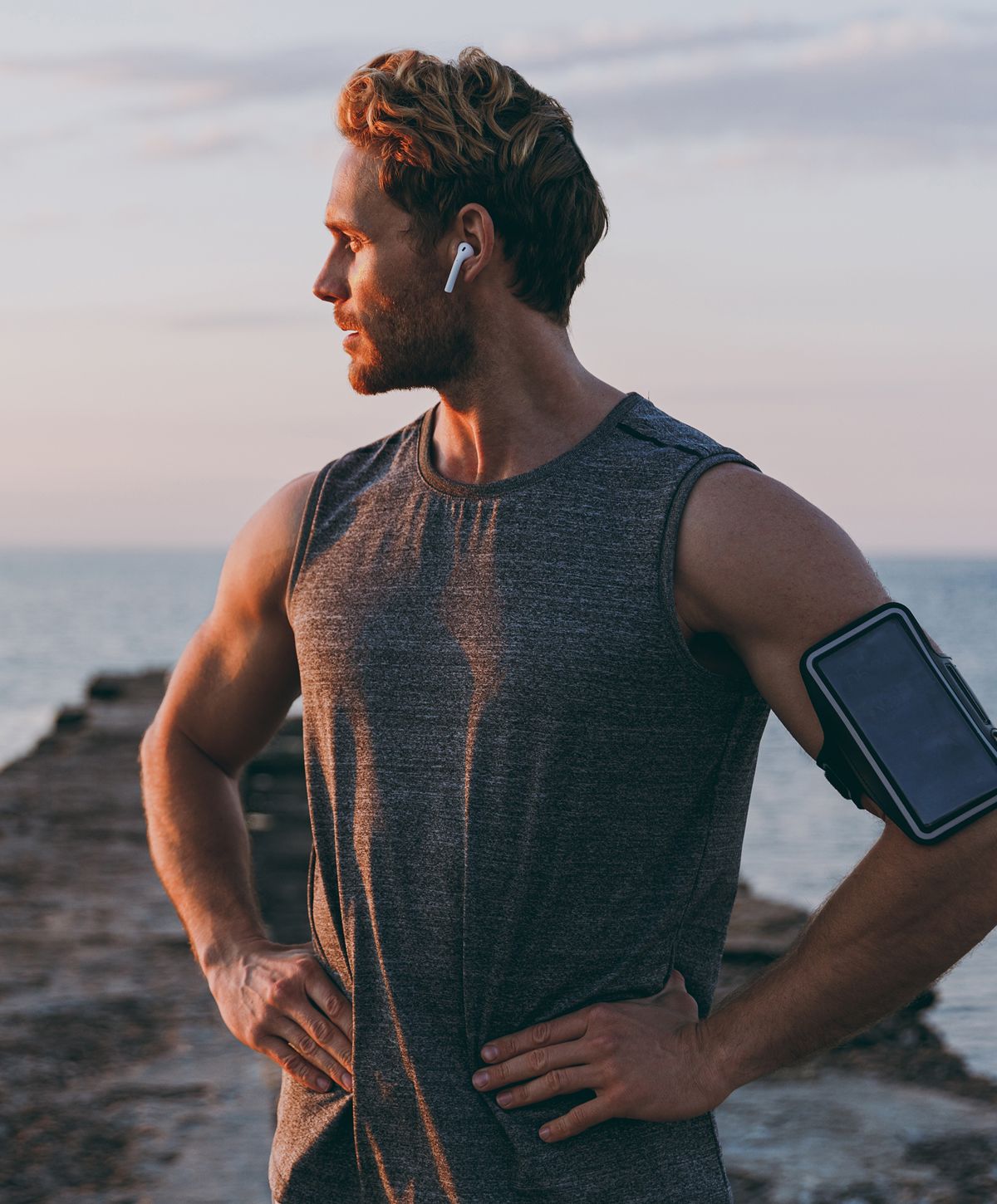 A man in a sleeveless gray athletic shirt stands on a rocky pier, looking off to the side. He wears wireless earbuds and has a phone armband on his left arm.