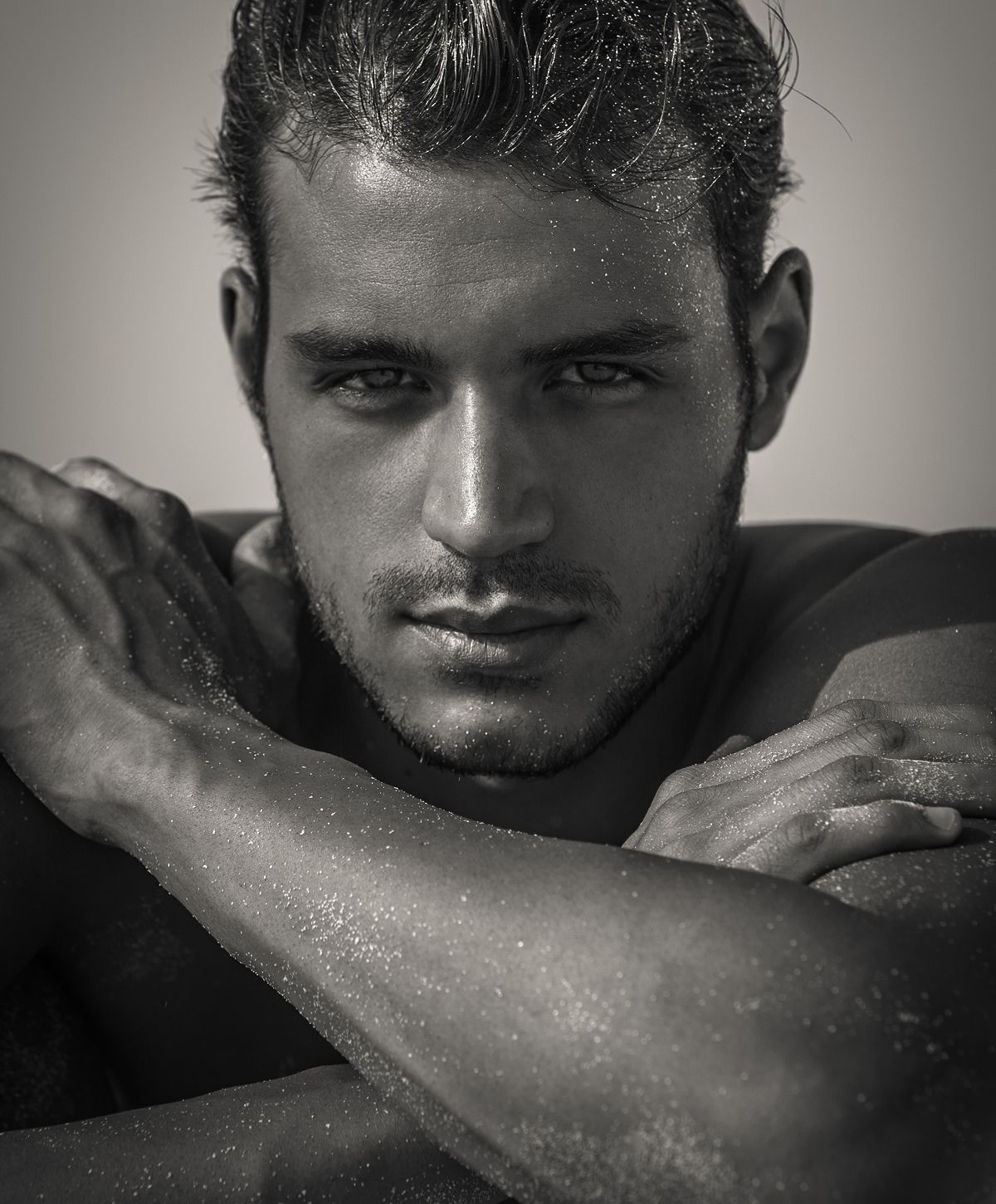 A close-up portrait of a young man with a strong jawline and intense gaze, resting his arms on his shoulders. The image emphasizes his facial features and wet, tousled hair, with a subtle sheen on his skin.