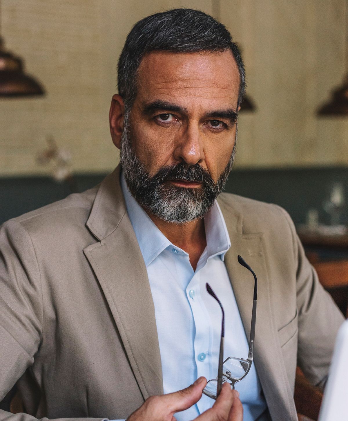 A man with a beard and short hair wearing a beige suit jacket and a light blue shirt, holding a pair of glasses in one hand and looking directly at the camera.