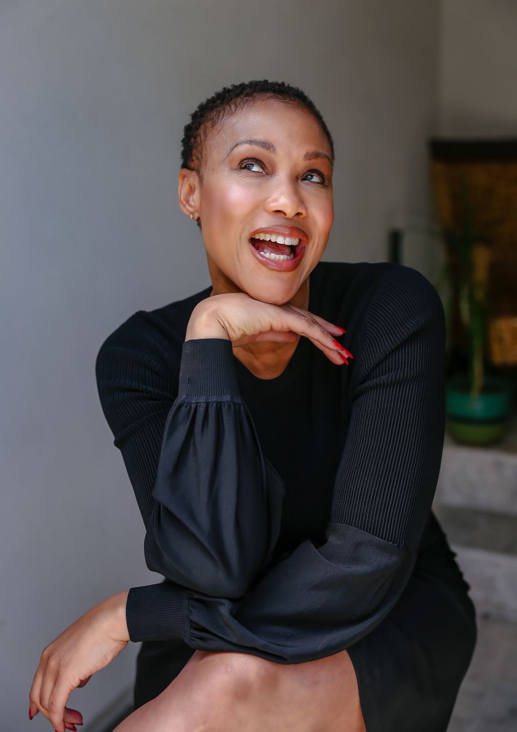 A woman with short, curly hair wearing a black long-sleeve dress, smiling and resting her chin on her hand.