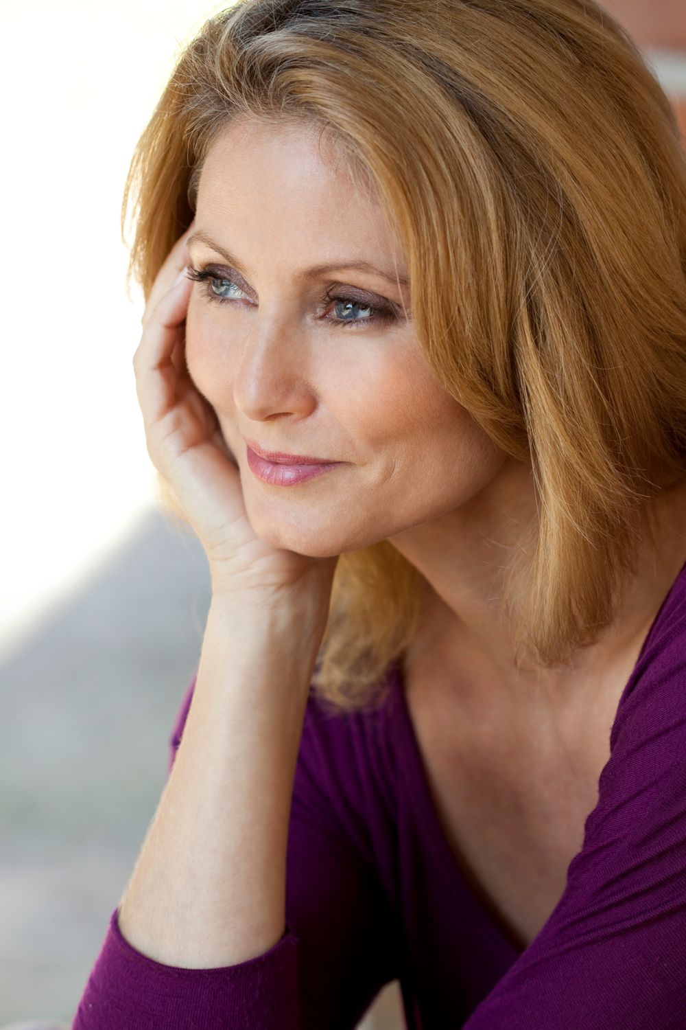 A woman with long, blonde hair, wearing a purple top, resting her chin on her hand and smiling softly, with a thoughtful expression.