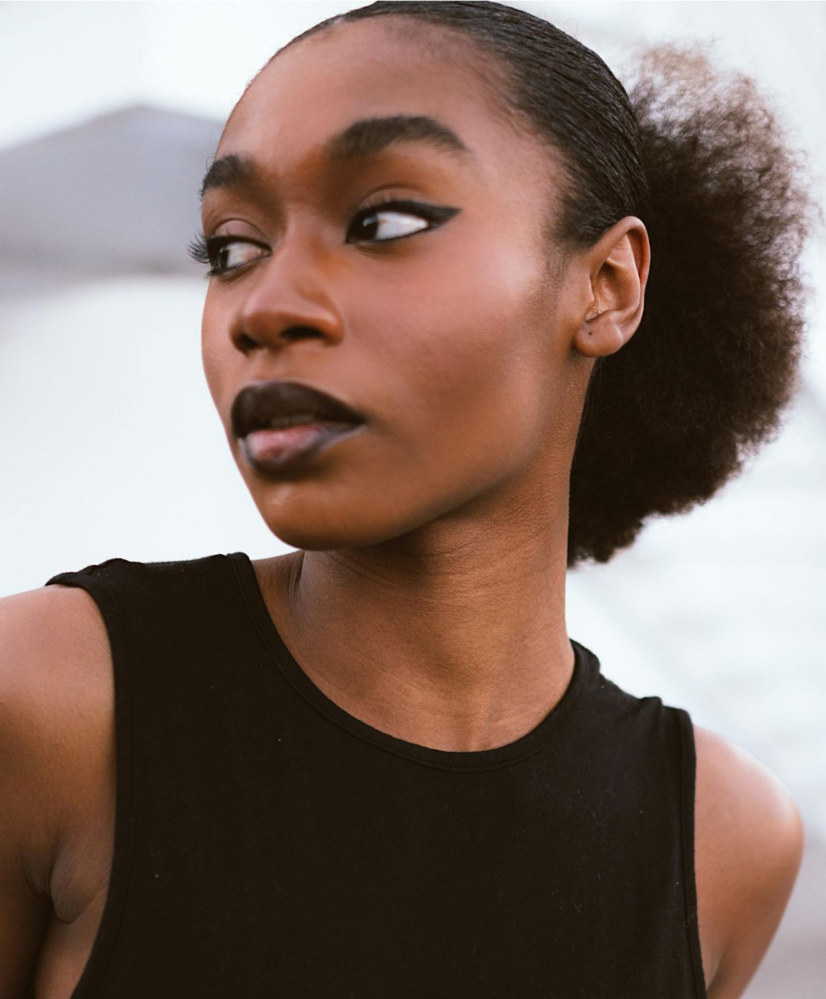 A woman with a sleek hairstyle wearing black lipstick and a sleeveless dark top, gazing over her shoulder with an expressive face.