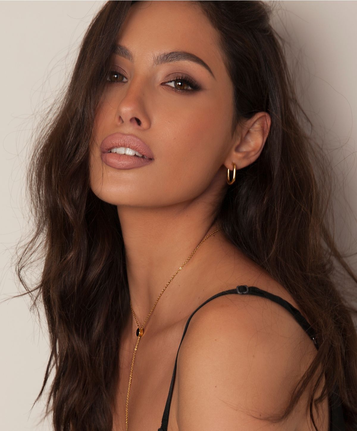 A close-up portrait of a woman with long, wavy dark hair, wearing a black top. She has a natural makeup look emphasizing her eyes and lips. She is adorned with a gold necklace featuring a small pendant and gold hoop earrings.