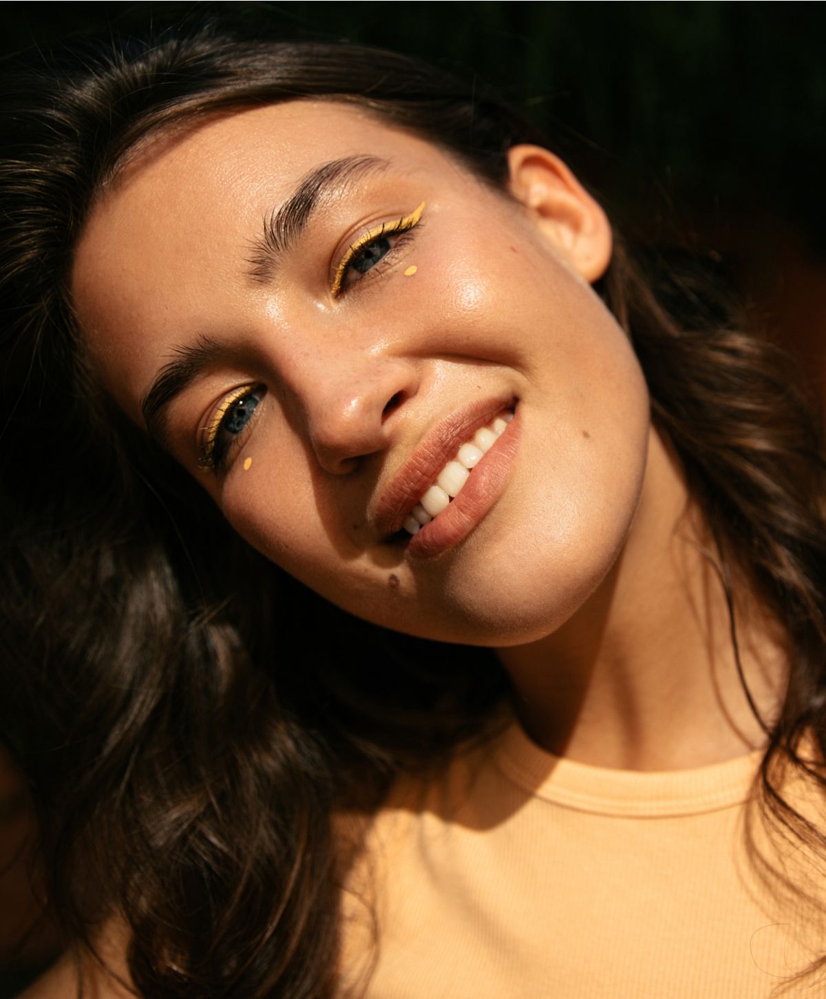 A woman smiling with wavy brown hair, wearing a light yellow top, and sporting bold yellow eyeliner and small yellow dots on her cheeks.