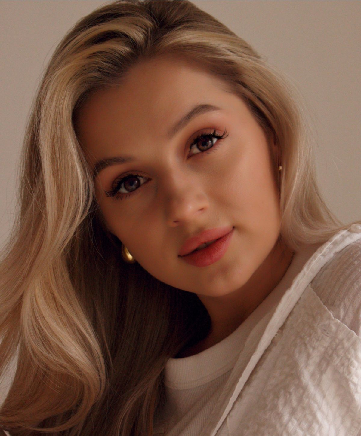 A young woman with long, wavy blonde hair, wearing a white top and gold hoop earrings, looking directly at the camera with a soft smile.