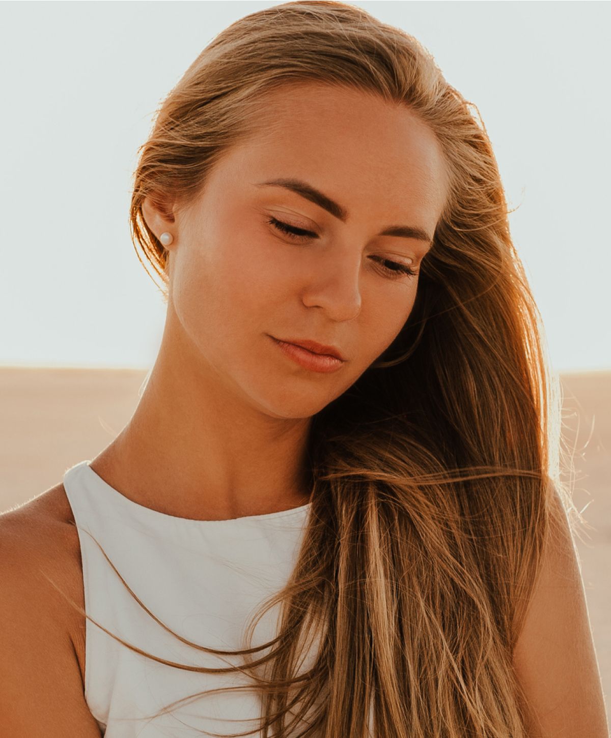 A woman with long, flowing hair, wearing a white top, looking down with a soft expression.