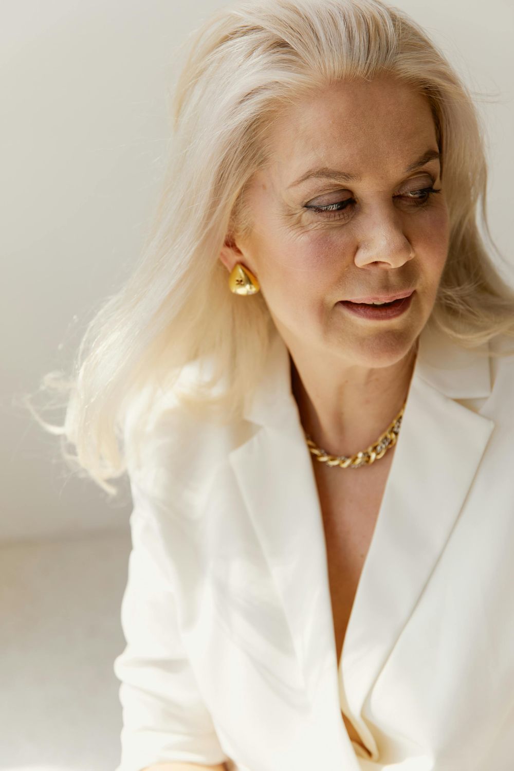 A woman with long, light blonde hair wearing a white blazer and a gold chain necklace, looking slightly to the side with a soft expression.