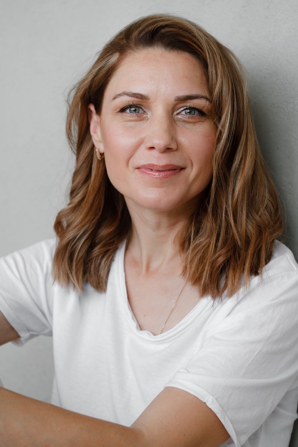 A woman with shoulder-length wavy brown hair, wearing a white t-shirt, smiling at the camera.