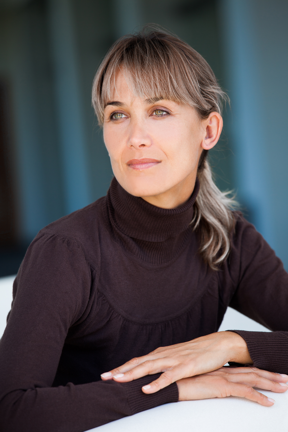 A woman with long, light brown hair pulled back, wearing a brown turtleneck sweater, looking thoughtfully to the side with a calm expression on her face.
