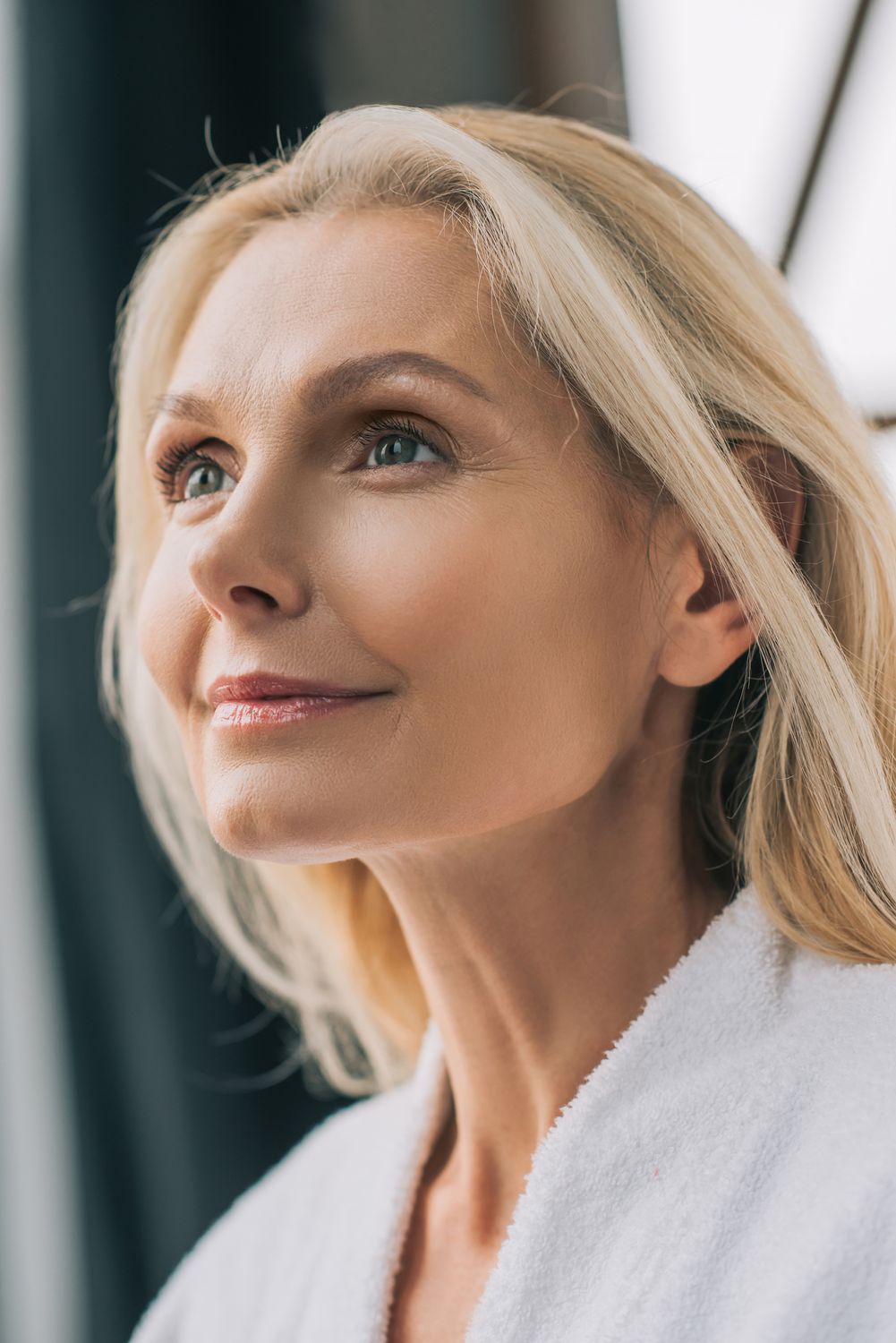 A close-up portrait of a woman with long blonde hair, looking upward with a serene expression. She has fair skin and is wearing a white robe.