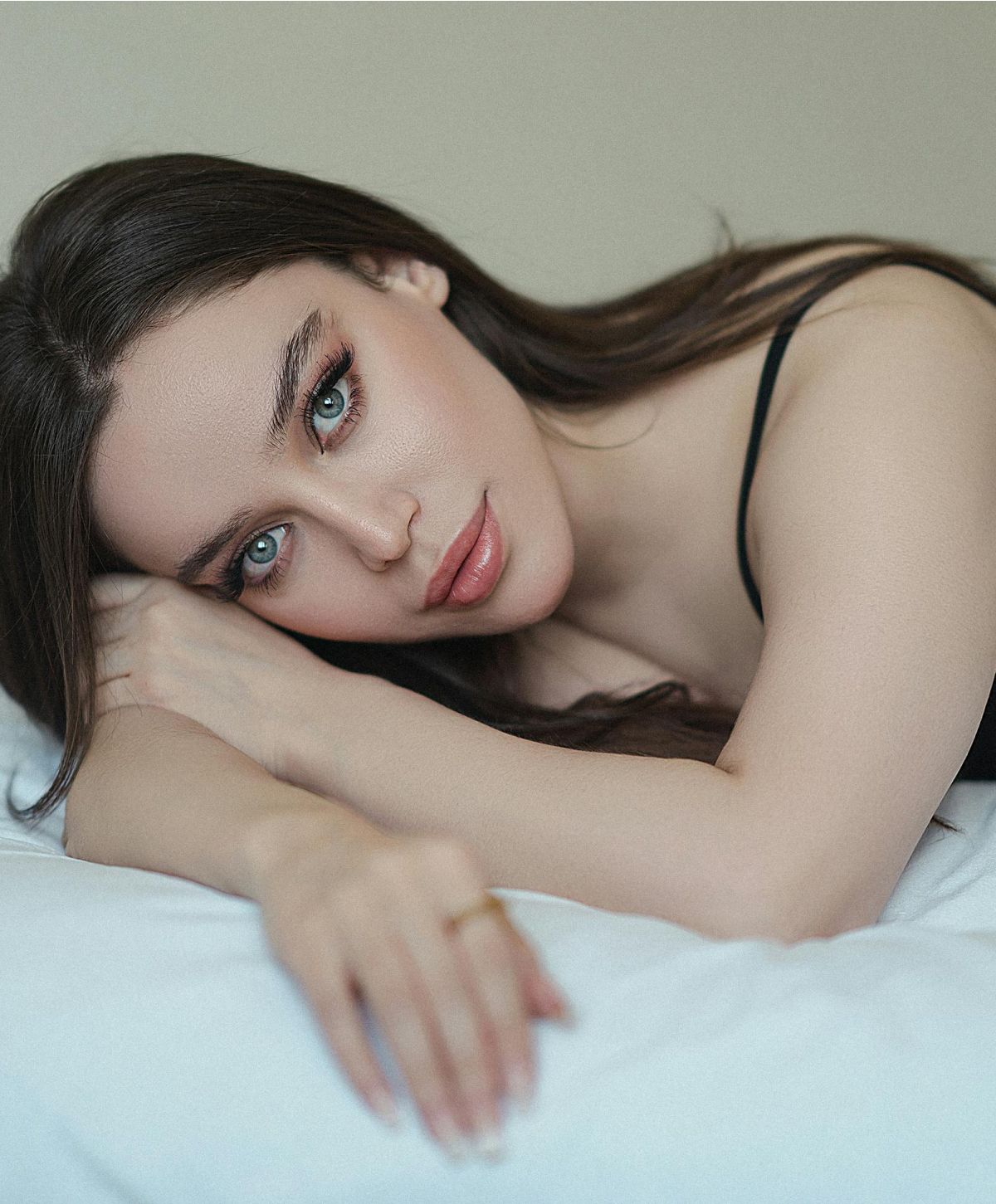 A woman with long brown hair resting her head on her hand, looking directly at the camera with striking blue eyes and natural makeup.