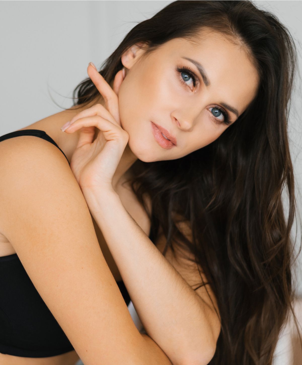 A woman with long dark hair poses with one hand touching her chin, wearing a black top, and looking thoughtfully at the camera.