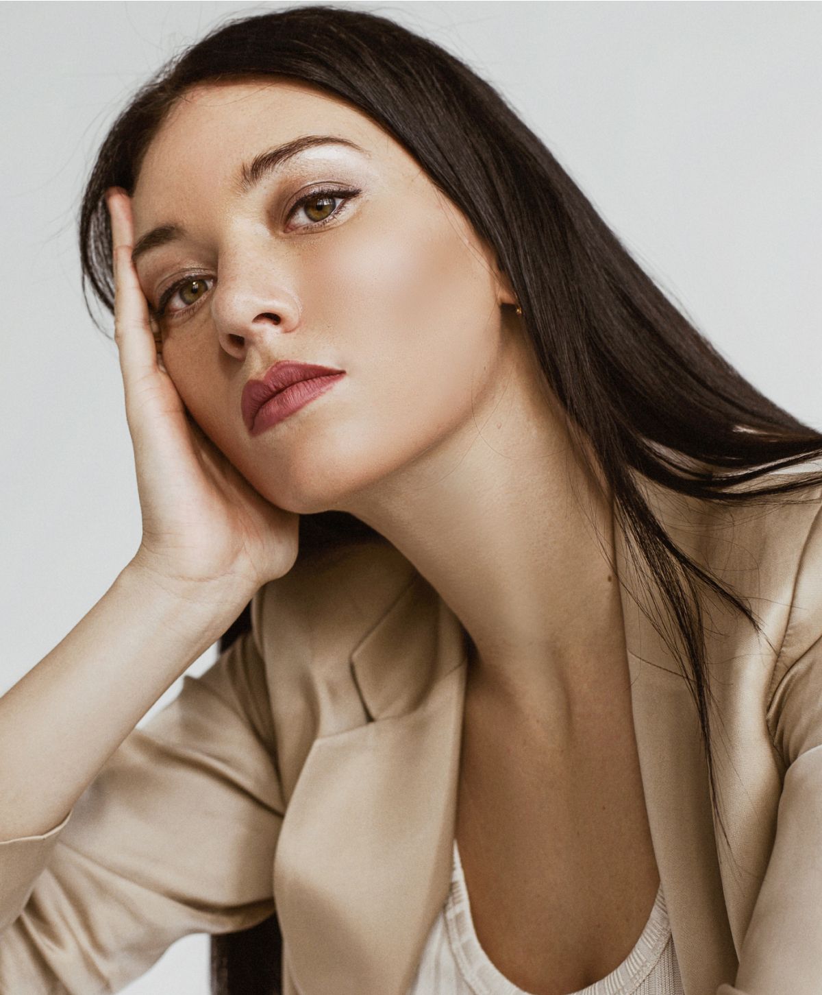 A woman with long dark hair, wearing a beige blazer, rests her head on her hand while looking thoughtfully at the camera. She has subtle makeup with a focus on her lips.