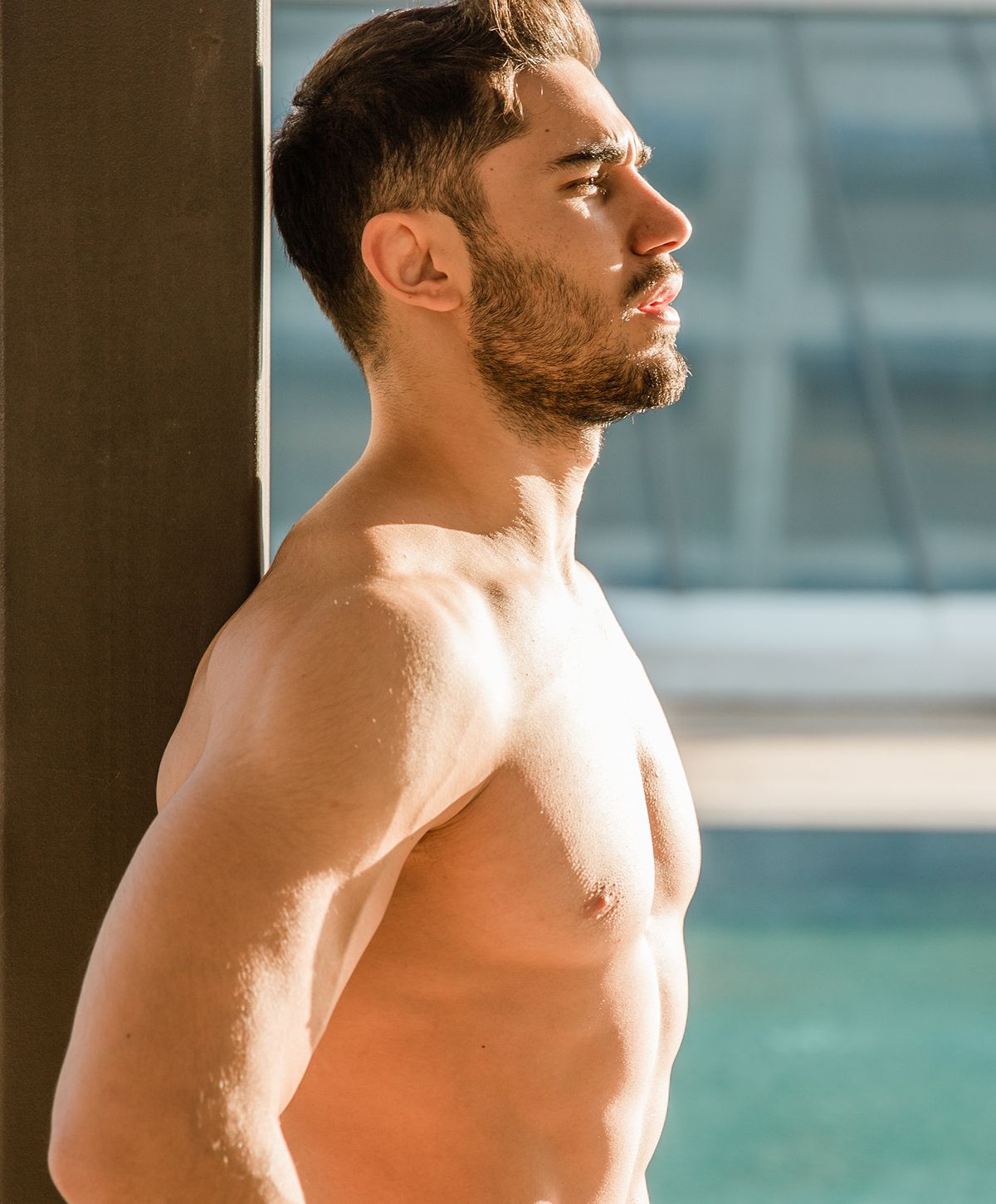 A man with a beard and short hair stands sideways, leaning against a vertical surface, looking upwards with a serene expression. His upper body is bare, showcasing his muscular physique.